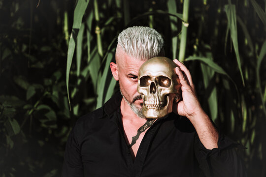 Brutal bearded mature male with tattooed chest holding human skull and looking at camera with challenge while standing against tall green plants