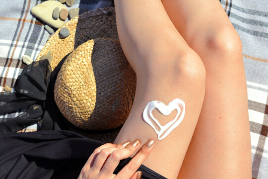 Woman applying suntan cream lotion, sunscreen cream in heart shape on her legs. Sun protection concept. Hat, body, summer, vacation.