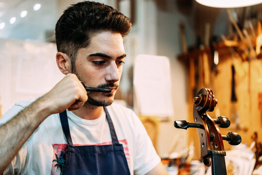 Serious repairman in apron standing with tuning peg and modern violin in workshop