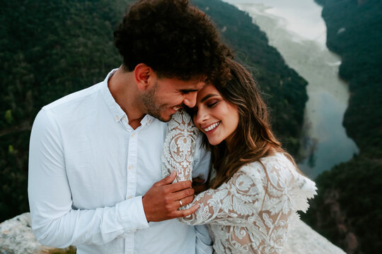 From above gentle young couple n stylish wedding outfits standing together with eyes closed on top of rock against amazing scenery of Morro de Labella in Spain
