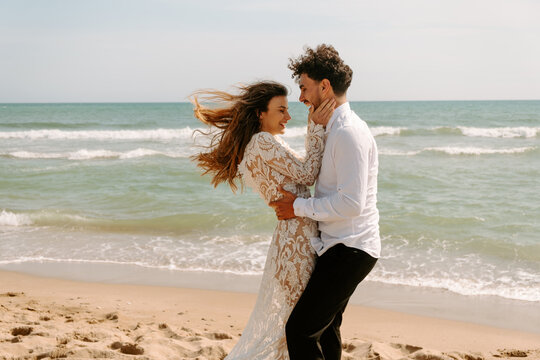Side view of cheerful young bride and groom in stylish wedding costumes standing embracing each other with eyes closed on sandy beach near waving sea and enjoying each other