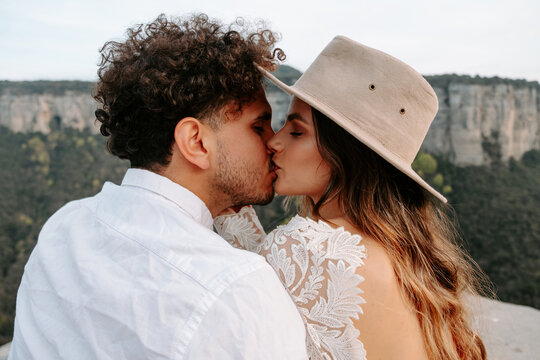 Back view of happy young couple in stylish wedding outfits kissing tenderly while enjoying romantic moments together against blurred mountainous landscape of Morro de Labella in Spain