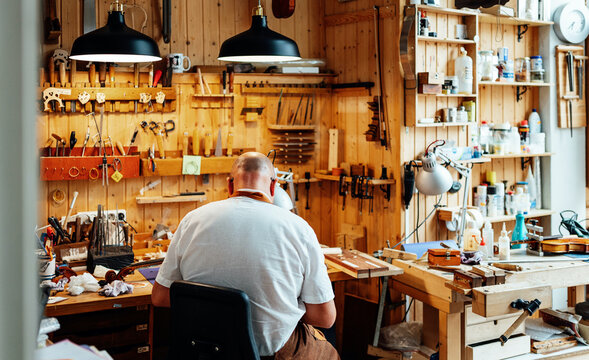 Back view of unrecognizable male luthier master sitting on chair and catching tool from wall while making instruments in workshop