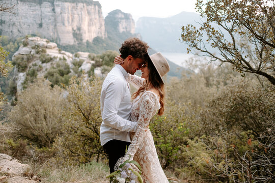 Side view of romantic young newly married couple in wedding outfits with bouquet embracing and kissing while standing against picturesque mountain landscape of Morro de Labella in Spain