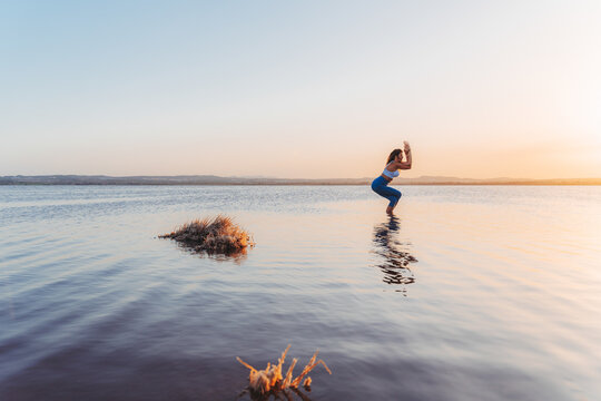 Full length side view of flexible slim female in activewear performing Garudasana pose while practicing yoga alone on lake shore in summer evening