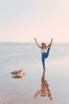 Full body of female in sportswear standing in lake water and performing standing split asana with arm raised while practicing yoga in sunny evening