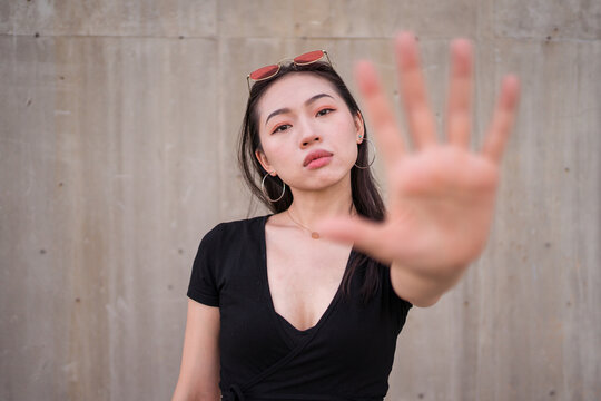 Young Asian female standing on street with outstretched arm and demonstrating stop sign with palm while looking at camera