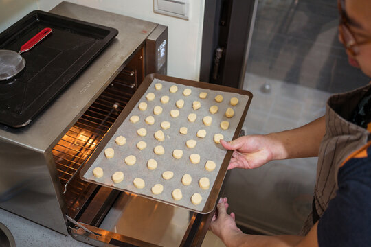 From above of crop baker in apron placing baking sheet with cookies into preheated oven at home