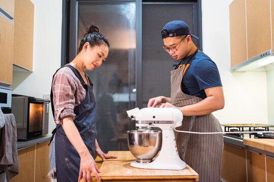 Side view of Asian man and woman in aprons watching stand mixer working process while preparing cream in kitchen
