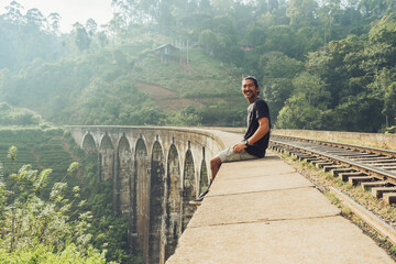 Side view of cheerful ethnic male tourist sitting on old stone viaduct Nine Arch Bridge with railroad leading through green jungle in Sri Lanka