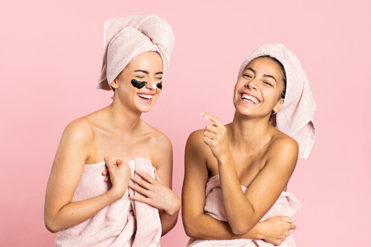 Cheerful young diverse females friend wrapped in bath towels smiling happily while enjoying spa charcoal treatment against pink background