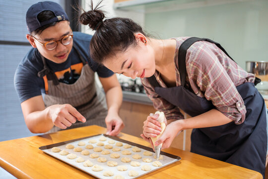 Asian guy helping smiling girlfriend with piping bag to pipe batter for pastries on baking pan at counter