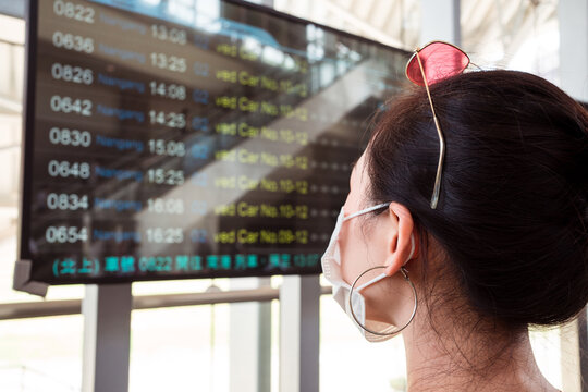 Back view of unrecognizable female in medical mask standing at railway station and looking at train schedule during coronavirus outbreak