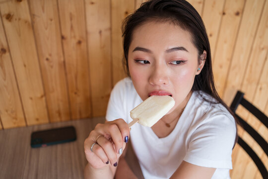From above ethnic female biting sweet ice cream and looking away while sitting at table in cafe
