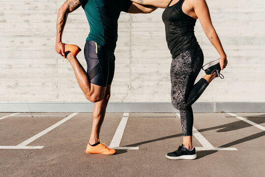 Cropped unrecognizable side view of strong sportsman and sportswoman leaning on each other and stretching legs before training in city on sunny day