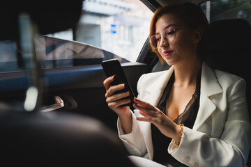 Determined female entrepreneur in elegant costume sitting in modern automobile and browsing mobile phone while checking messages and working remotely