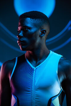 Serious concentrated young African American male athlete standing against blurred blue neon background in dark studio looking away