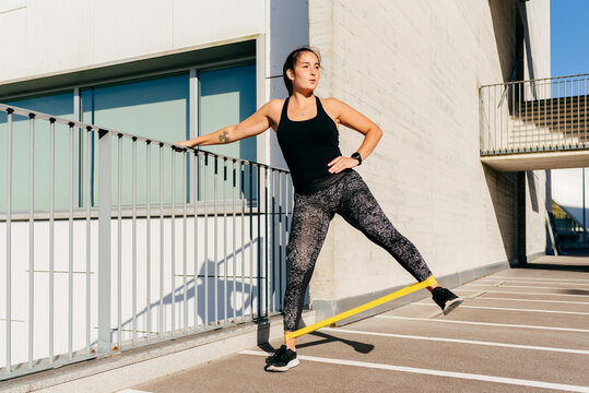 Slim female athlete in sportswear doing exercises with elastic band while standing near metal fence during workout in city and looking away