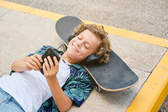 Boy with printed shirt lying on the floor with his head on a skateboard listening to music on his mobile