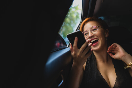 Cheerful female entrepreneur sitting in luxury car and recording voice message while working and using cellphone