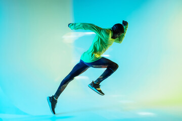 Full body side view of young African American male runner in colorful tracksuit sprinting from start position in studio with bright neon illumination