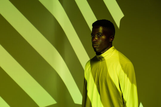 Young African American male athlete in sportswear looking away while standing under green neon illumination near corner with striped shadows in studio