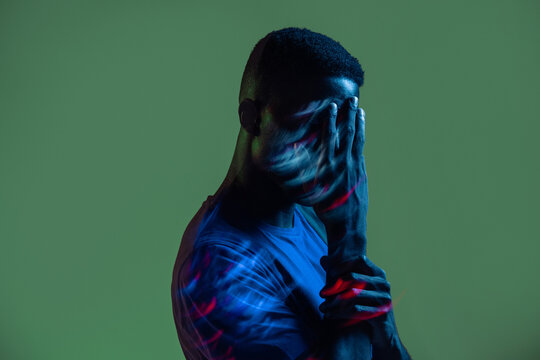 Serious confident young African American male with colorful neon lights on face looking at camera against green background