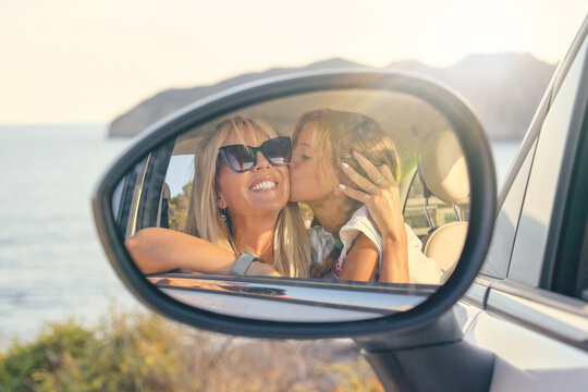 Car mirror with the image of a blond daughter kissing and hugging her mother and looking at the mirror in front of the sea