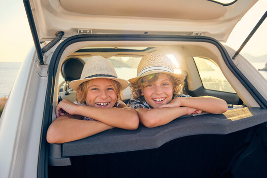 Two children in straw hats leaning in the back seats of a car sticking their heads out of the trunk with an expression of happiness