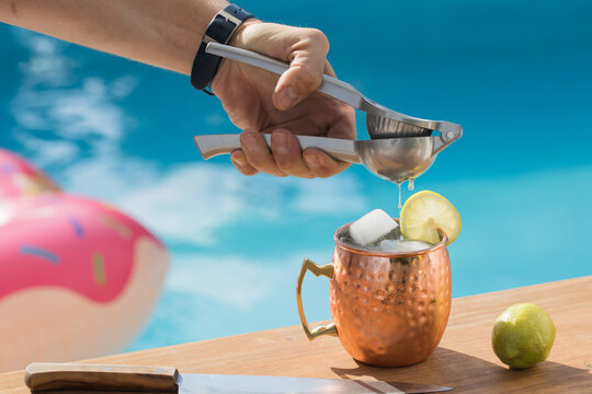 Crop anonymous man with squeezer adding lime juice into copper cup with Moscow mule cocktail near swimming pool in summer day