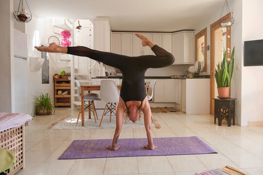 Young blonde woman doing a variation of the yoga handstands position on top of a mattress in the kitchen of a modern house