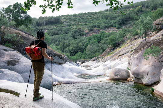 Back view of anonymous male traveler with backpack and trekking sticks standing on rock and enjoying picturesque view of river in highland area