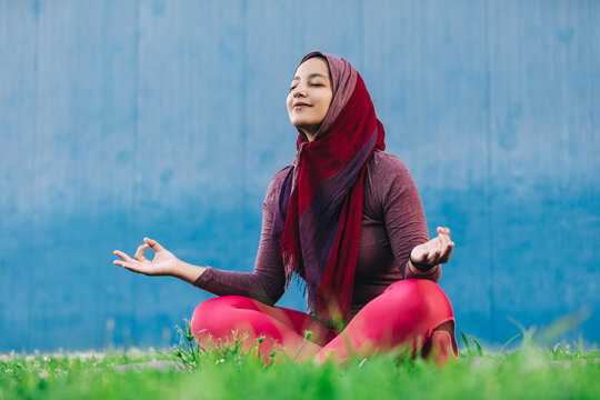 Serene Arab female in sportswear and hijab sitting on mat on lotus pose with eyes closed in park and doing yoga in Padmasana with closed eyes - Powered by Adobe