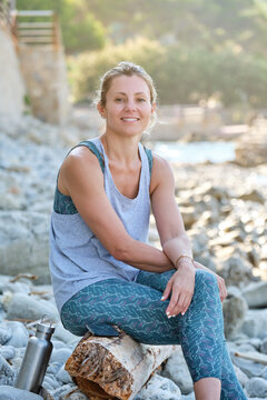 Smiling female in sportswear sitting on tree trunk while resting after meditation on rocky seashore and looking at camera