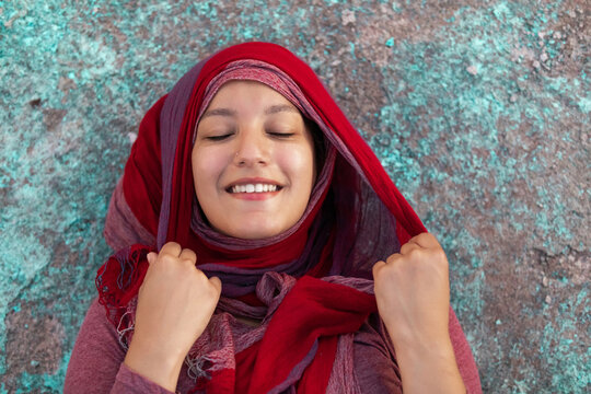 Happy Arab female wearing hijab leaning on concrete colorful building in city smiling with eyes closed