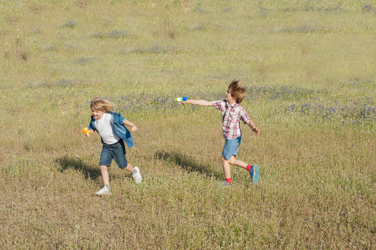 Positive brothers in summer wear running in meadow and playing with water guns while entertaining during weekend in countryside