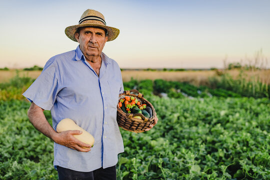 Aged male farmer standing barefoot with wicker basket of fresh fruits and vegetables on garden bed and looking at camera