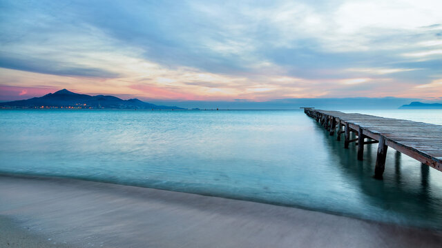 Majestic scenery of wooden quay leading to calm sea water under sunset sky in Playa de Alcudia in long exposure