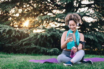 Full body cheerful young curly haired female in sportswear with headphones sitting on grass and browsing smartphone while resting with bottle of water after workout in summer park