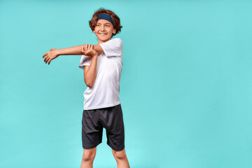 Happy teenage boy in sportswear looking away and smiling while stretching his arms before workout or training, standing isolated over blue background © Kostiantyn