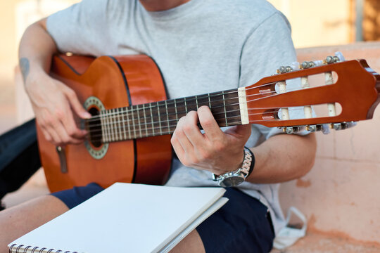 Creative male musician sitting on stone bench on street and reading notes while preparing for playing guitar