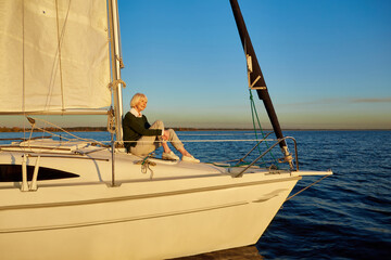 Enjoying luxury life. Beautiful senior woman sitting on the side of sailboat or yacht deck floating in the calm blue sea at sunset, looking away and smiling