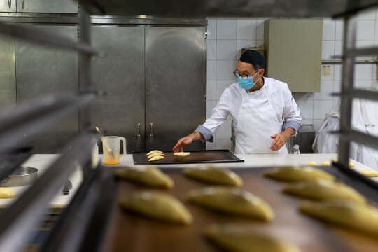 Senior male chef in medical mask and apron preparing pasty in kitchen in bakery during COVID 19
