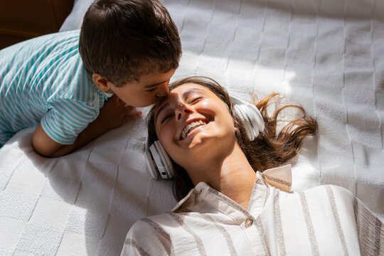 High angle view of son kissing his mother's forehead listening to music while lying on bed