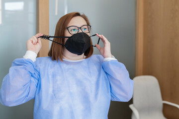 Female medical worker in blue uniform and eyeglasses putting on black protective mask with adjustable strap while working in modern clinic during coronavirus pandemic