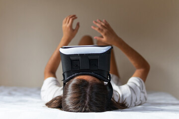 Happy young female in VR headset while lying on bed and exploring cyberspace at home