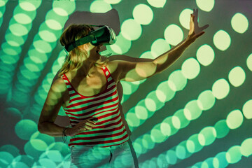 Unrecognizable young female in casual wear and VR headset getting new experience and touching virtual object in room with colorful projector illumination