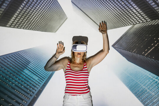 Unrecognizable female in casual wear and VR headset standing with arms raised against contemporary skyscrapers inside virtual city
