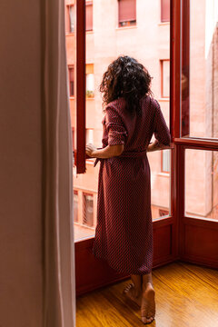 Back view of unrecognizable thoughtful woman looking out to big windows of her apartment