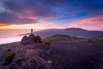 Back view of anonymous male traveler standing with outstretched arms on rocky hill in highlands and enjoying amazing view of clouds